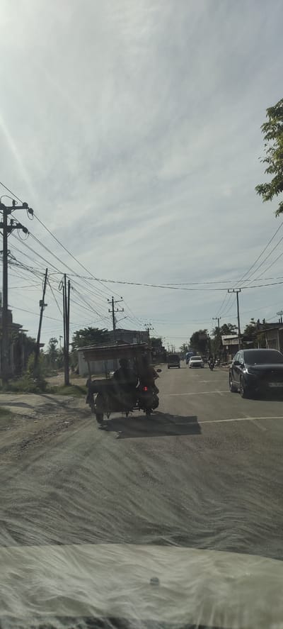 The image captures a street scene in Beurandang, Indonesia. A three-wheeled motorcycle with a covered sidecar is crossing an intersection. Several other vehicles are visible: a dark-colored SUV to the right, a white sedan, and a motorcycle. Overhead, electrical wires stretch across the sky. The road shows signs of wear. A portion of the dashboard is visible at the bottom of the frame.