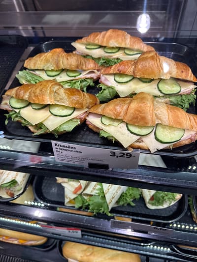 A close-up, slightly angled, medium shot displays a selection of food items within a refrigerated display case. The main subject is a tray filled with croissant sandwiches, sliced in half and filled with lettuce, ham, cheese, and cucumber slices. To the right, other containers are visible, holding tortilla wraps and sandwiches with bread. A price tag is placed on the tray with the croissant sandwiches, reading "3,29-" and bearing text in Latvian, including the words "Kruasāns" (croissant), "ar šķiņķi un sieru" (with ham and cheese), "SVAIGS GURĶIS, SALĀTI" (fresh cucumber, lettuce), and the company "Circle K Latvija". The text on the price tag suggests the food is sold by Circle K in Latvia. The location of Saldus is not mentioned in the image.