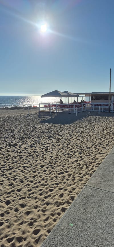 The image depicts a sunny beach scene in Vila do Conde, Portugal, with a cafe or restaurant situated on the sand near the water. The foreground is dominated by rippled sand, suggesting it is a popular spot. A paved walkway runs along the right side of the frame. In the middle ground, a white-railed structure houses tables and chairs under large, light-colored umbrellas. Several people are seated at the tables, enjoying the view of the ocean. The sea is calm and reflects the bright sunlight, with a clear blue sky above. A single pole stands to the right of the cafe, and some rocks are visible along the shoreline. The overall atmosphere is relaxed and inviting, characteristic of a beachside establishment.