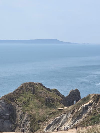 This image shows Durdle Door, a natural limestone arch on the Jurassic Coast near the village of West Lulworth, Dorset, England. The scene is dominated by the vast blue sea, with a hazy distant coastline visible on the horizon. In the foreground, the rugged, grassy cliffs rise dramatically from the water. A prominent natural rock arch, Durdle Door, is visible in the mid-ground, with a small opening through its center. A wooden viewing platform is situated on the cliff face near the arch. A winding staircase, made of stone steps, ascends the hillside on the right side of the image, indicating a popular hiking route. Several people are scattered across the landscape, appearing as small figures walking on the paths, enjoying the scenic view. The overall impression is a natural coastal landscape with evidence of human activity through the pathways and visitors.