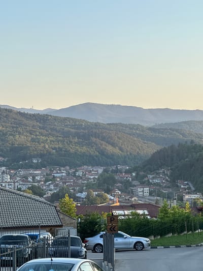 This is a vertical, eye-level, outdoor shot of a town nestled in a valley surrounded by verdant, rolling hills under a clear, pale blue sky. In the foreground, a paved road curves to the right, bordered by a black and white curb and a metal fence. Several cars are parked and visible: a silver sedan is in the middle of the frame, facing right, with a wooden signpost for parking next to it. Behind it, a darker colored fence encloses more parked cars, including a dark gray Porsche. To the left of the fence, the grey tiled roof of a building is prominent.

In the midground, the town of Kerenite, Bulgaria, unfolds with a dense cluster of buildings featuring red-tiled roofs, interspersed with greenery. The buildings are of varying sizes and architectural styles, indicative of a residential area.

The background is dominated by layered hills covered in dense, dark green forests. The distant hills fade into a soft, hazy blue, suggesting atmospheric perspective. On the crest of the furthest hill to the left, a tall, slender structure, possibly a tower or monument, is visible against the sky. The lighting suggests it is late afternoon or early evening, with a soft, even illumination across the scene. There are no visible people in the image.