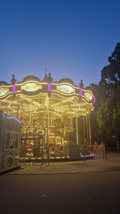 A brightly lit carousel, adorned with numerous golden lights and purple accents, stands in a park at dusk. The carousel features ornate decorations and painted panels depicting landscapes. To the left of the carousel, a ticket booth labeled "KASSZA" is visible. The ground in front of the carousel is paved with cobblestones. In the background, trees and a few people are present. The sky is a deep blue, indicating it is twilight. The scene is located in Keszthely, Hungary.