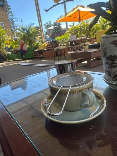 This image depicts an outdoor cafe scene in Bellingen, Australia, bathed in bright sunlight. In the foreground, a glass-topped table holds a ceramic coffee cup and saucer, with a metal tea strainer resting inside the cup. To the left of the cup, a small metal pot with a long handle is visible. The table's surface reflects the surrounding environment, including trees and sky. In the background, there is outdoor seating made of pallets and cushions, an orange umbrella, and lush green plants. A woman in a red scarf is seated on a green sofa in the middle ground. The overall setting suggests a relaxed cafe atmosphere.