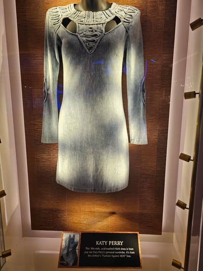 The image displays a dress, presumably showcased in a museum or exhibit, located in Hollywood Seminole Reservation, United States of America. The dress, made of acid-washed denim, features a form-fitting design with unique cutout details along the neckline and sleeves. It is mounted on a mannequin and positioned against a wall with a textured, bronze-colored finish. To the right of the dress, a placard bearing the title "KATY PERRY" provides information about the garment. The text on the placard states that the dress is from Katy Perry's personal wardrobe and is part of H&M's "Fashion Against AIDS" line. The scene is illuminated by overhead spotlights, casting shadows and highlighting the textures of the dress and the wall.