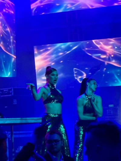In this image, two women are performing on a stage with a futuristic, glowing background. The woman on the left is wearing a sequined black halter top and sparkly green pants, with her hair pulled back in a ponytail. She is gesturing with her left hand. The woman on the right is also wearing a similar sequined outfit and has her hair in a ponytail. Both women appear to be singing or dancing. In the foreground, parts of the audience are visible, with one man in a suit and glasses in sharp focus. The stage is lit with blue and purple lights, and there are some sound equipment cases behind the performers. The image does not provide any specific details about the location being the city of Akhalubani, Georgia.
