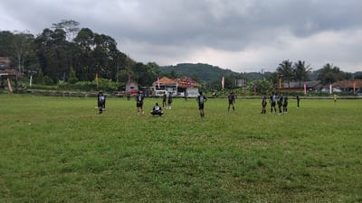 This image captures an outdoor scene on a grassy sports field in Kecamatan Panjalu, Indonesia, under an overcast sky. A group of young men, likely a sports team, are gathered on the field. They are dressed in matching dark-colored jerseys, primarily black, with white numbers visible on their backs, such as "13" and "55". One player wearing jersey number 13 is crouched down in the center-left foreground, while others stand in various positions around the field, suggesting a break in play, a training session, or a pre-game huddle.

In the background, traditional houses with distinct reddish-brown tiled roofs and white walls are visible, interspersed among dense green trees and lush vegetation. Beyond these structures, rolling hills or mountains are discernible, indicating a scenic, natural landscape typical of rural Indonesia. Along the edge of the field near the houses, colorful flags or banners in shades of pink, purple, and yellow/orange add decorative elements. Another player in a bright yellow jersey is visible further to the right in the background. The cloudy sky suggests a daytime setting, possibly late morning or afternoon, without direct sunlight. The scene depicts a community sports event or practice session taking place in a verdant, rural Indonesian environment.