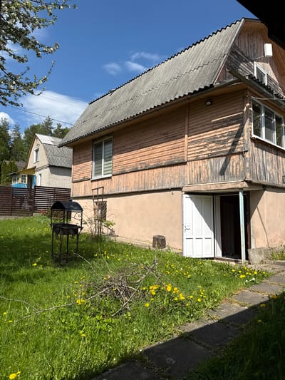The image depicts a two-story wooden house in Пятрышкаўскі сельскі Савет, Belarus. The house has peeling paint on its exterior and a grey slate roof. A partially open white door leads into what appears to be a basement or ground floor entrance. The house is surrounded by a lawn with dandelions. A grill stands on the lawn. In the background, part of another building with a blue accent can be seen. The sky is blue with scattered clouds.