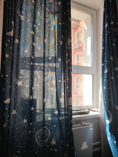 In the city of Putilkovo, Russia, sunlight streams through a window, illuminating a scene featuring blue curtains with a star and moon pattern. The curtains are drawn back to reveal a view of a building with orange and white facades, featuring multiple windows and balconies. On the windowsill, a small keyboard, a pen, and a magazine are visible. Below the windowsill, a white radiator with a mesh cover is present. The overall impression is a cozy indoor scene juxtaposed with an urban exterior.