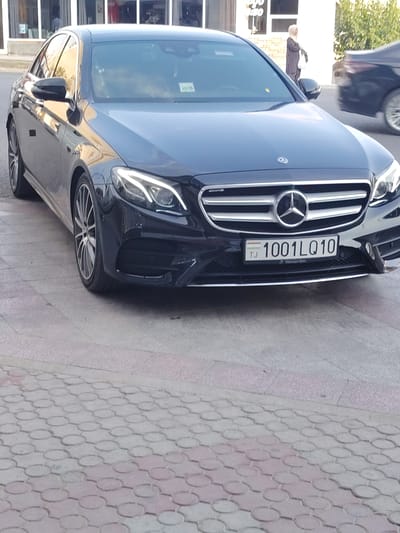 A black Mercedes-Benz sedan is parked on a street with a patterned pavement. The car is the primary subject, with its front grille, headlights, and license plate clearly visible. The license plate reads "1001LQ10 TJ" and has a small symbol of the flag of Tajikistan on the left. Below the license plate, the text "Mercedes-Benz" is visible.
In the background, a woman in a patterned dress and hijab is standing near a building with large windows. Another dark-colored car is partially visible in motion to the right of the Mercedes. The scene appears to be outdoors during daylight. The presence of the Tajikistan flag on the license plate and the general setting suggest the location is Khujand, Tajikistan. The weather appears to be clear.