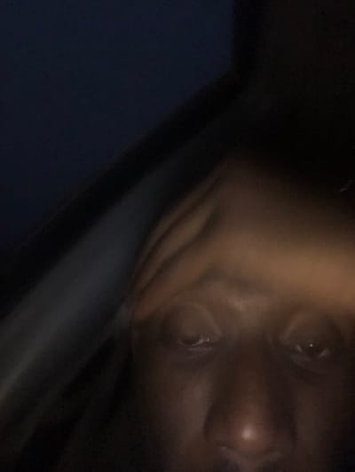 A close-up, dimly lit image focuses on a person's face. The skin appears smooth, with visible features like the nose, eyes, and subtle creases. The background is dark, suggesting a low-light environment. To the left of the face is a vertical, dark gray element, possibly part of the environment or a structural component. The overall composition draws attention to the face and its features.