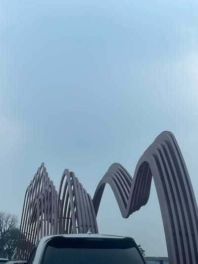 The image depicts a modern outdoor sculpture with a wavy, segmented design, likely made of metal, against a hazy, pale blue sky. The sculpture dominates the upper half of the frame. In the foreground, the rear of a white SUV is visible, with its roof and darkened rear window occupying the bottom portion of the image. To the left of the sculpture, bare trees are visible, suggesting a cooler season or climate. The overall impression is one of an artistic installation in an urban or suburban setting, possibly a park or public space. There are no people visible in the image. The location is specified as Cijantra, Indonesia.