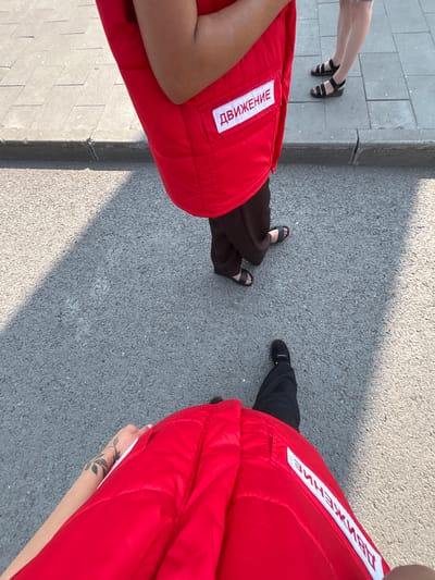 The image shows a high-angle, slightly rotated view of two people standing on an asphalt surface, likely a sidewalk or street. Both individuals are wearing bright red vests or jackets with white patches containing Cyrillic text. The text on the vest on the left reads "ДВИЖЕНИЕ" (DVIZHENIE), meaning "Movement" or "Action" in Russian. The text on the vest on the right is partially visible but appears to be the same.

On the left side of the frame, a person's legs are visible, wearing black sandals with ankle straps, and dark pants. Their left foot is closer to the camera, showing a pedicure. The background on the left consists of light-colored paving stones, indicating a walkway.

On the right side of the frame, the legs of another person are visible, wearing black trousers and dark shoes. A tattoo of a vine with leaves is visible on the forearm of the person wearing the red vest on the right.

The scene is outdoors, and the presence of shadows suggests it is daytime, likely sunny. The asphalt texture and paving stones provide context for an urban or semi-urban outdoor setting. The activity depicted is simply standing, suggesting a pause or waiting moment. There are no overt expressions of emotion, but the positioning and attire might hint at participation in an event or work related to the "Movement" indicated by the text. The location context provided is Belovo, Russia.
