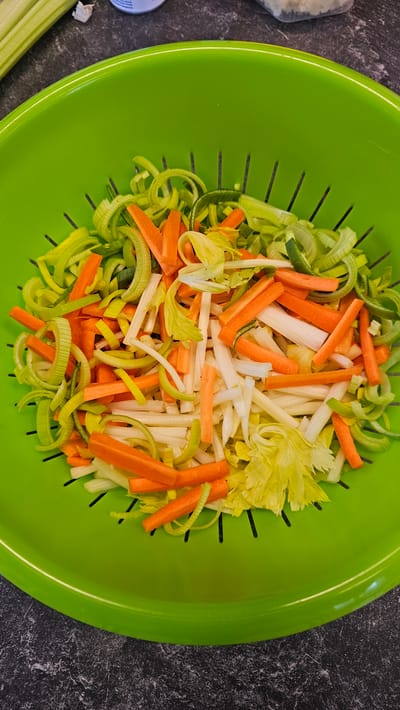 An overhead shot captures a vibrant green colander filled with an assortment of freshly chopped vegetables, resting on a dark gray, speckled countertop. The colander's inner surface features small, elongated slots. The main subjects are the mixed vegetables, meticulously cut into uniform pieces.
Dominating the mix are bright orange carrot sticks, appearing julienned or in small batonnets. Interspersed among these are light green and white rings or half-rings of what appears to be sliced leeks, and pale white, thin stalks of celery, some still retaining small, yellow-green celery leaves. The various colors of the vegetables create a visually appealing contrast within the green colander.
The activity depicted is the preparation of ingredients, as the vegetables are clean and uniformly chopped, suggesting they are ready for cooking or washing. To the top-left of the colander, a blurry, light-colored object, possibly a container, is partially visible. In the bottom-left corner, a single, unchopped stalk of celery can be seen, partially off-frame. No visible text is present in the image. The lighting is bright and even, illuminating the scene clearly.