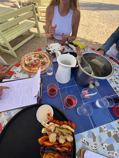 An outdoor dining scene is depicted in the Ramat Negev Regional Council, Israel, featuring three individuals seated at a rectangular mosaic-tiled table. The table is covered with blue, white, and patterned tiles. A person with long blonde hair, wearing a white sleeveless top, is seated at the top-center, with their forearms resting on the table; a ring is visible on the left ring finger and a smartwatch on the left wrist. To the left, a person's hand rests on an open menu attached to a clipboard. To the right, the arm of a person wearing a denim garment is partially visible near a silver ice bucket containing an uncorked bottle of white wine and ice.
Food items on the table include a round pizza topped with mushrooms, basil, and cheese, resting on a wooden board. A ceramic bowl contains a dark dish covered with white crumbled cheese. A black serving tray holds a platter of roasted vegetables, specifically carrots, cauliflower, sweet potatoes, and bell peppers, alongside a small white bowl of hummus garnished with paprika.
Beverages and related items include a white ceramic pitcher, one clear glass filled with water, two stemless glasses containing a pink liquid, and two empty clear glasses. A small clear container holds multiple white sugar packets. A set of two shakers, likely salt and pepper, are also present. A yellow object is visible on a white napkin near the person in the white top. The background features light-colored wooden benches and a ground surface composed of gravel or crushed earth, indicating an outdoor, sunny environment.