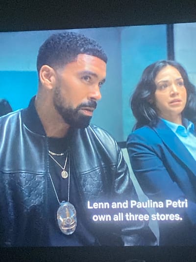 The image, originally rotated, displays two individuals—a man in the foreground and a woman behind him—in what appears to be an indoor setting with a plain light blue background.
The man, positioned on the left side of the frame, has dark, short, curly hair and a neatly trimmed beard. He is wearing a black, possibly leather, bomber-style jacket with a subtle, embossed pattern, resembling interlocking motifs (e.g., Gs) across the fabric. Underneath, he wears a dark shirt. He is adorned with two necklaces: a shorter one with a small circular gold-colored pendant, and a longer one featuring a larger, clear, oval-shaped pendant that encases a metallic element or design. His expression is serious, and he appears to be looking towards the woman.
The woman, partially visible in the upper right background, has dark hair styled around her shoulders. She is dressed in a light blue collared shirt beneath a dark blue blazer or jacket. Her gaze is directed towards the man, also with a serious or attentive expression.
The scene appears to be a still from a television show or video, capturing a moment of interaction or dialogue between the two characters.
Notable details include the distinctive patterned jacket worn by the man and the prominent necklaces. Overlaid on the image, mostly across the man's shoulder and jacket, is white text that reads: "Lenn and Paulina Petri own all three stores."
There are no visual cues within the image to specifically indicate the location is Gosa, Nigeria. The lighting suggests an artificial, possibly studio, environment.