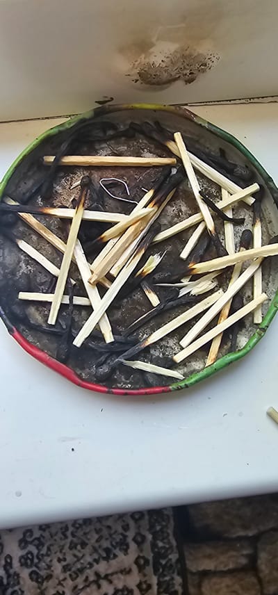 The image shows a pile of burnt and unburnt matchsticks in a circular metal container. The container has a green and red rim. The surface below the container is white, and the background shows a patterned cloth and brickwork. There are no people visible in the image. The scene appears to be indoors or on a protected surface.