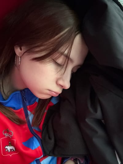 A close-up shot depicts a person sleeping. The person has fair skin, brown hair with bangs partially covering their face, and is wearing hoop earrings. They are dressed in a red and blue zippered jacket featuring a crest-like design with a white bear. A dark, seemingly heavy garment or blanket is draped over the person. The background is obscured, focusing attention on the sleeping individual. The location of Kukushtan, Russia, is not discernible from the visual information provided.