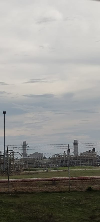The image depicts an industrial complex, likely a power plant or refinery, under an overcast sky. In the foreground, a chain-link fence topped with barbed wire separates the viewer from the industrial site. Beyond the fence, various structures, including tall cylindrical smokestacks and interconnected piping systems, are visible. Some buildings with grid-like facades are also present. The ground within the fenced area appears to be a mix of dry vegetation and some green grass. A single tall lamp post with a square head stands to the left of the frame. The overall impression is one of a functional, industrial landscape. The location in the city of Glokaron, Indonesia, suggests this is a significant industrial facility within that region.