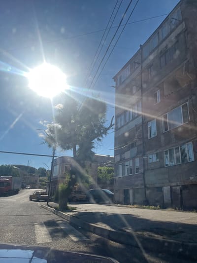 This is a photograph taken in Gladzor, Armenia, on a sunny day. The image is oriented vertically, with the sun prominently featured in the lower left corner, casting lens flares and bright light across the scene. The sky is a clear blue, with a few wispy clouds.

The foreground is dominated by a tree and power lines, with the sun's glare making it difficult to discern finer details. To the right, a sidewalk and a street are visible, with shadows stretching across them, indicating it is likely mid-day. The street is paved with asphalt and appears to be a two-lane road.

In the background, several buildings are visible. On the left, a multi-story, aged apartment building with multiple windows stretches across the frame. Its facade appears weathered. Further back and to the right, other buildings are present, some with visible windows and balconies. One building appears to have a modern facade with large windows. A few vehicles are parked along the street, including what looks like a dark SUV and a yellow vehicle. The overall setting suggests an urban or semi-urban environment, possibly residential or mixed-use. The bright sunlight and clear sky suggest pleasant weather. No people are clearly visible in the image. There is no discernible text.