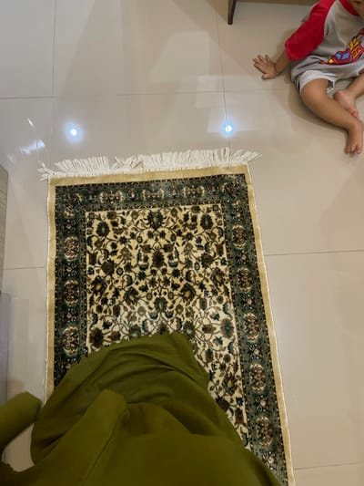 This overhead shot, taken in Ngasinan, Indonesia, captures an indoor scene featuring a small child, a partial view of an adult, and a patterned rug on a tiled floor.

In the upper left quadrant of the image, a young child, likely a toddler, is lying on their back or side on light-colored, square tiled flooring. They are dressed in a light grey t-shirt with red sleeves and white shorts or underwear. Their bare legs and feet are visible, along with one arm and hand. The child appears relaxed, resting or playing on the floor.

Dominating the center and right side of the frame is a rectangular prayer mat or small rug. The rug features an intricate floral design with a cream-colored main field, bordered by patterns in dark green and brown, accented with gold details. One end of the rug displays short, white fringes.

To the far right, next to the rug, a person is partially visible, clad in olive green clothing, possibly a long dress or robe. Only a section of their lower body and draped fabric can be seen, suggesting they are sitting or kneeling beside the rug, perhaps in preparation for or during prayer.

The floor is a light beige or cream tiled surface, reflecting bright overhead light. The overall setting is a domestic interior, quiet and well-lit. No text is visible in the image.