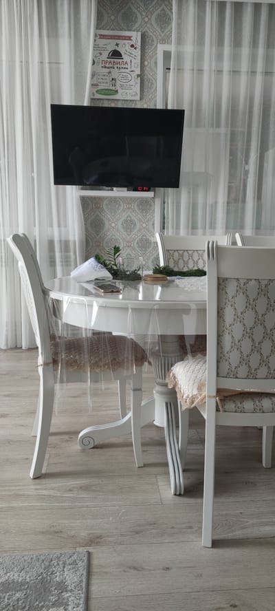 A dining set with white chairs and a round table covered with a transparent plastic tablecloth is positioned in a room. On the table, there is a white napkin, a clear glass vase with greenery, a mobile phone, and a wooden coaster. The table is supported by a white pedestal base. The chairs have upholstered seat cushions with a light-colored pattern. Behind the table, a wall is decorated with a light green and gray patterned wallpaper. A flat-screen television is mounted on the wall above the wallpaper. To the left and right of the television are sheer white curtains. The floor is covered with light-colored laminate flooring, and a portion of a gray rug is visible in the lower left corner. The scene appears to be indoors, likely in a dining area, in Икское Устье, Russia.