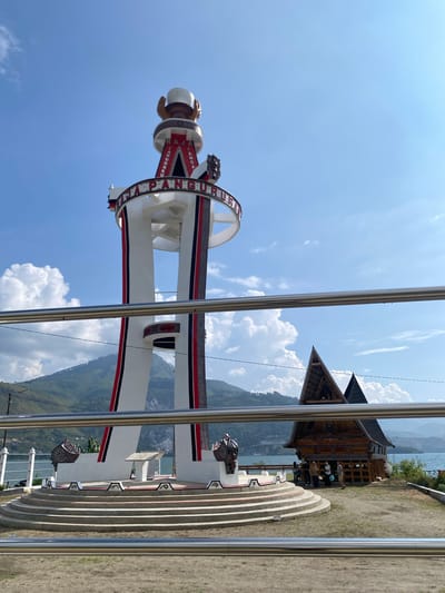The image shows a monument in the city of Parlondut, Indonesia. The monument is a tall, white structure with red and black accents, topped with a large, white sphere held by two hands. A circular sign around the monument reads "RAJA PARANGURUN." The monument is surrounded by tiered steps. To the right of the monument is a traditional wooden house with a steep, triangular roof. In the background, there is a body of water, lush green hills, and a clear blue sky with white clouds. Several people are visible near the wooden house. A metal railing runs across the foreground of the image, partially obscuring the view.