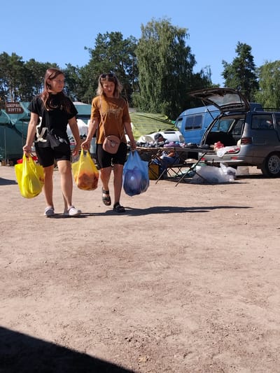 In Irshansk, Ukraine, two young women are walking outdoors, each carrying shopping bags. The woman on the left, with brown hair, wears a black t-shirt and black shorts, and carries a yellow shopping bag in her right hand. The woman on the right, with blonde hair, wears a tan t-shirt with a leopard print and black shorts, and carries a yellow shopping bag in her left hand and a blue shopping bag in her right. Both women appear to be in a parking lot or market area, with tents and vehicles in the background. The ground is dirt and gravel, and the sky is clear and blue.