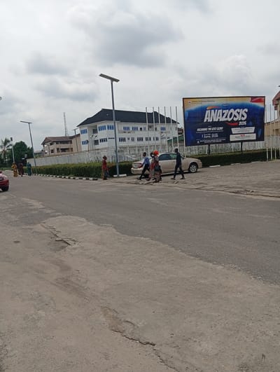 The image depicts a street scene in Elimgbu, Nigeria, with a large billboard dominating the background. The billboard advertises an event called "ANAZOSIS 2026" presented by Deeper Life Campus Fellowship, Rivers Central. It states the theme as "REGAINING THE LOST GLORY" and is targeted towards students, corps members, staff, and campus dwellers. The event is scheduled for Thursday, June 18th to Sunday, June 21st, 2026, and will take place at the Deeper Life Camp Grounds, East-West Road, Rumuodara, Port Harcourt. The billboard also mentions "Life-transforming Messages, Spiritual Seminars, Counselling and Healing, Relationship and Networking Sessions" and lists "Pastor Pius Idume" as the preacher.

In the foreground, a road with visible wear and tear is visible. Several individuals are crossing the street, and a few cars are parked or driving on the road. The environment appears to be an urban setting with a building complex and a white fence visible behind the road. The sky is overcast with clouds.