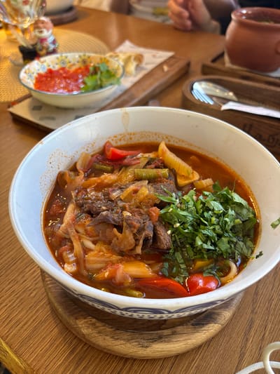A top-down, slightly angled shot captures a bowl of hearty stew, likely a traditional dish from Solnechnoye, Russia, placed on a wooden table. The stew is rich with chunks of tender meat, visible green beans, and pieces of red bell pepper, all swimming in a savory, reddish-brown broth. The dish is generously garnished with finely chopped fresh green herbs, adding a vibrant contrast. The bowl itself is white with a decorative blue and white pattern along its rim, and it rests on a round, wooden serving board.
To the left of the main dish, a small, decorative ceramic bowl holds a bright red puree, possibly a sauce or relish, topped with a sprig of green herb. This smaller bowl also has a colorful, floral pattern. In the background, glimpses of other tableware and a hand reaching for something suggest a meal is in progress. The setting appears to be a restaurant or dining area, evidenced by the wooden table and cutlery holder in the upper left corner, which contains a spoon and fork. The lighting suggests it might be daytime. The overall impression is of a satisfying, home-style meal being enjoyed in a casual dining environment.