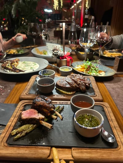 A table laden with culinary delights: grilled steak, lamb chops with asparagus, french fries, and salads, each meticulously presented on stylish plates and wooden boards. Accompanying sauces in small bowls enhance the flavors. Glasses of wine hint at a sophisticated dining experience, suggesting a luxurious restaurant setting with a warm and inviting ambiance.