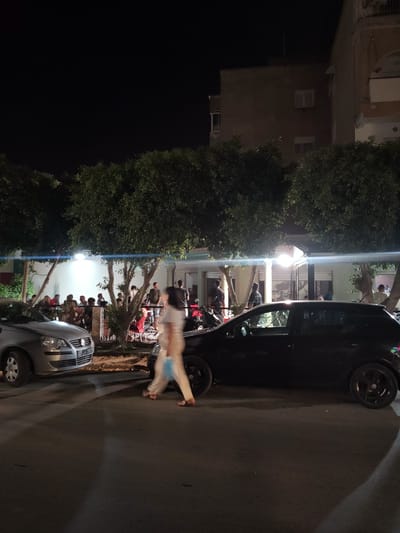At night, a street scene is depicted in an urban environment in Tunisia. A silver Volkswagen Polo is parked on the left side of the road. A black hatchback car is parked to its right. In the background, a group of people are gathered outside a brightly lit establishment, possibly a restaurant or cafe, shaded by trees. A person is walking in the foreground, blurred due to motion. Buildings are visible behind the trees. The road surface is asphalt, and shadows are cast by the cars and trees.