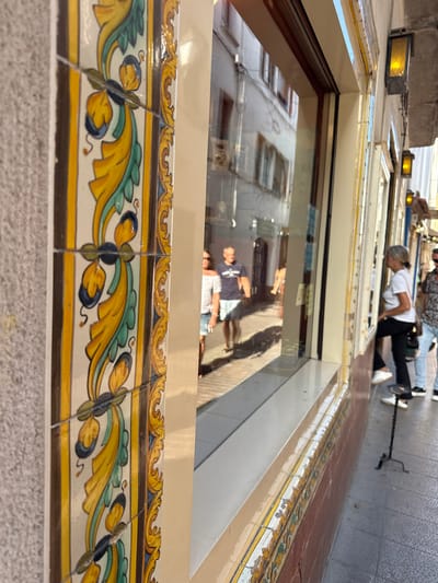 The image shows a reflection in a large window, revealing people and buildings. The reflection is distorted and upside down, suggesting the photo was taken from a low angle looking upwards. In the foreground, decorative tiles are visible, featuring a yellow, green, and blue floral pattern. These tiles appear to be on the exterior of a building.
The reflection shows several individuals. One person, in the reflected image, is wearing a white t-shirt and black pants, and is in a bent-over position, possibly looking into something. Another person in the reflection is wearing a dark t-shirt and shorts. Beyond them, more people and the facades of buildings are visible, suggesting an urban or town setting. The lighting in the reflection suggests it is daytime. The presence of decorative tiles and the architecture hint at a location in Spain, consistent with the provided context of Tossa De Mar. The overall scene appears to be a street view captured through a reflective surface, with the focus on the details of the building's exterior and the inverted reflections of the activity happening in front of it.