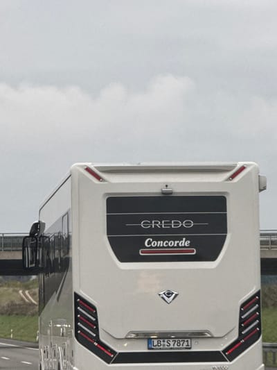 This image depicts the rear of a white Concorde Credo motorhome, bearing German license plate LB S 7871, on a paved road identified as being in Dornstadt, Germany. The vehicle displays "CREDO" and "Concorde" branding on its black rear panel, positioned above a silver trim piece and the license plate. A rear-view camera is mounted centrally above the branding. Red marker lights are visible on the top rear corners of the vehicle. On the lower rear, vertical taillight clusters incorporate red and white segmented lights. A large side mirror assembly is visible on the motorhome's left side. The environment is characterized by an overcast sky with light gray clouds. In the background, a concrete bridge structure crosses horizontally above the road. To the left, a green grassy embankment with a white road marker is visible. No individuals are present in the scene.