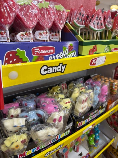 This image depicts a retail display of various confectionery items, likely in a store in Acıpayam, Turkey. The foreground features a close-up of individually wrapped marshmallow candies, shaped like bunnies and other figures in pastel colors such as pink, yellow, and blue. These are arranged in black plastic trays. Above these, a prominent yellow shelf band runs across the image, displaying the word "Candy" in large black lettering, with a smaller inscription "Marshmallow Handmade" visible on the packaging below.

Further up, a display of lollipops is visible. On the left, several strawberry-shaped lollipops are presented in clear plastic wrappers, with small labels indicating "Candy Pop's" and featuring strawberry imagery. To their right, a cluster of watermelon-slice-shaped lollipops, also individually wrapped, is arranged in a cardboard display box. This box also features graphics of watermelon slices and a cartoonish green character. A price tag indicating "Fe 80 TL" is affixed to the yellow shelf band. The overall environment suggests a candy aisle or section within a shop.