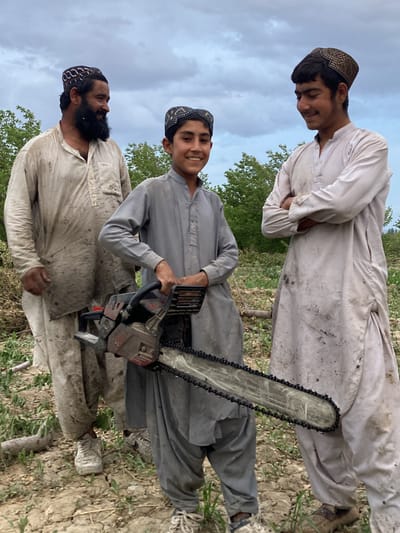 The image displays three males of varying ages posing together outdoors under an overcast sky. From left to right, there is an adult man, a young boy, and a young man, all wearing traditional long-sleeved tunics, likely shalwar kameez tops, which are visibly soiled with dirt or mud. Each person also wears a distinct patterned cap.

The adult man on the left has a dark beard and smiles as he looks to his right. The central figure is a young boy, likely a pre-teen, who holds a chainsaw with both hands. He beams directly at the camera, showcasing a cheerful expression. The chainsaw itself appears robust, with a long bar and chain. On the right, a young man, possibly a teenager or young adult, stands with his arms crossed, looking to his left with a gentle smile.

The setting appears to be a rural or semi-agricultural area. The ground is dry and uneven, covered with sparse green plants and scattered pieces of wood or branches, suggesting recent land clearing or tree felling activity. Lush green trees form the background, further emphasizing the outdoor, natural environment. The soiled clothing and the presence of the chainsaw indicate that the individuals may have been engaged in manual labor related to forestry or land management. All three individuals display positive emotions, smiling for the photograph despite the evident work. No discernible text is visible on any object or clothing.