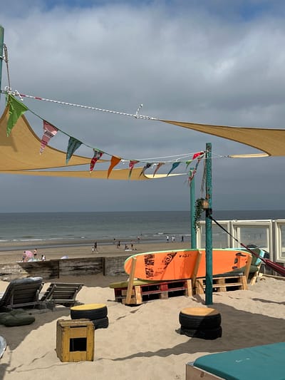 This is a medium shot of a beachside cafe in Zandvoort, Netherlands, on a cloudy day. In the foreground, sand-covered seating areas are arranged around a turquoise blue lounge chair. Two wooden sun loungers are placed on the sand to the left. A wooden crate with a square opening serves as a small table, and a black tire stacked with another tire forms a taller seating option to its right.
Behind the seating, a light orange surfboard stands upright, propped against wooden pallets. A green potted plant hangs from a teal-colored pole supporting a large tan sunshade that covers part of the scene. Festive pennant banners in various patterns and colors are strung across the sunshade.
In the midground, the sandy beach slopes down to the ocean, where gentle waves roll onto the shore. Several people are visible on the beach, some walking and others relaxing. A wooden barrier separates the cafe area from the beach.
In the background, the sea meets a hazy sky with scattered clouds. To the right, a section of a building with glass windows is visible. The overall atmosphere is relaxed and summery.