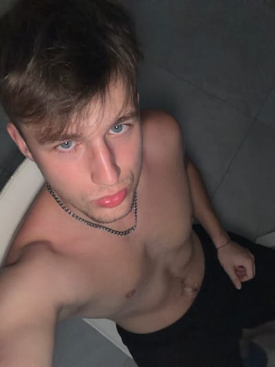 The image is a close-up, high-angle selfie of a young, shirtless Caucasian male with light brown hair and blue eyes. He is wearing a silver chain necklace and black pants. His skin appears flushed, and he has a neutral expression with slightly parted lips. The background is a dimly lit, tiled surface that appears to be a floor. There is a white chair partially visible on the left side of the frame. The setting does not provide any specific clues to identify a location in Vanino, Russia.