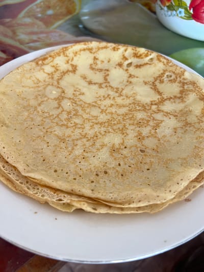 A close-up shot shows a stack of golden-brown crepes on a white plate. The crepes have a slightly uneven texture with small holes and darker brown lacy patterns, indicating they were likely cooked on a griddle. The plate itself is white with a thin silver rim, and it rests on a dark, textured surface, possibly a wooden table. In the background, a blurred image of a kitchen setting is visible, with a colorful bowl adorned with floral patterns and sliced oranges. The lighting is natural and bright, highlighting the appetizing appearance of the crepes. There are no people or animals present in the image. The setting appears to be domestic, likely a kitchen or dining area in a home.