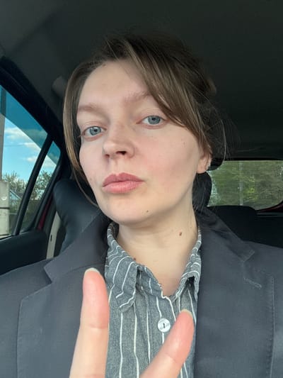 This image captures a close-up of a person, likely a young adult female, seated inside a car in Aksay, Kazakhstan. The person has fair skin, light brown hair parted on the side, and clear blue eyes. Her lips are adorned with light pink lipstick, and a small mole is visible on her neck. She is wearing a dark, possibly black, blazer or jacket over a grey and white striped collared shirt, which features a single white button near the neckline.

The person's right hand is raised towards the viewer, with the index and middle fingers extended to form a "V" sign, commonly known as a peace sign. Her expression is neutral as she looks directly at the camera.

The background shows the interior of the car, with dark upholstery visible. Through the car window on the left side of the frame, a bright sky with scattered clouds and green foliage, suggestive of trees or bushes, can be seen, indicating an outdoor, possibly rural or suburban setting during daytime with clear weather. The image is rotated counter-clockwise, causing the subject and background to appear tilted. There is no visible text in the image.