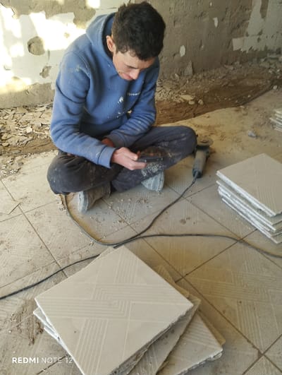 An indoor scene in Nurli zamin, Uzbekistan, depicts a male individual sitting cross-legged on a dusty, light-colored tiled floor. The individual is wearing a blue hooded sweatshirt, black trousers, and light-colored shoes, with dust visible on their clothing. Their dark hair is short. The individual holds a dark-colored smartphone in both hands and looks down at the screen. To the right of the individual, a grey and black angle grinder with a black power cord is visible on the floor. Several stacks of light-colored, square tiles featuring a geometric pattern are arranged on the floor in the foreground and mid-ground. The background reveals unfinished or partially demolished walls, with exposed plaster and debris accumulated along the base. The floor throughout the scene is covered with dust and small fragments of material.