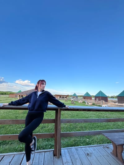 A young woman with auburn hair pulled back in a ponytail leans against a wooden railing. She is wearing a dark blue quarter-zip sweatshirt over a white t-shirt and black leggings. Her feet are clad in black sneakers with white laces. She is looking up and to her left, with a neutral expression.

The setting appears to be outdoors, possibly a rural area, given the presence of several small, wooden buildings with green, gabled roofs in the background. The buildings are arranged in a row, and beyond them, a fence encloses a grassy area where a few white goats can be seen grazing. The ground is covered in lush green grass dotted with small white flowers.

The sky is a clear, vibrant blue, with a few wispy white clouds present. The lighting suggests it is daytime, likely late afternoon or early evening, given the soft light. The overall atmosphere is peaceful and serene.

The image is oriented vertically, with the woman positioned in the right half of the frame, leaning against the railing. The railing is made of weathered wood and runs horizontally across the foreground. Beyond the railing, the grass and buildings recede into the distance. There is no visible text in the image.