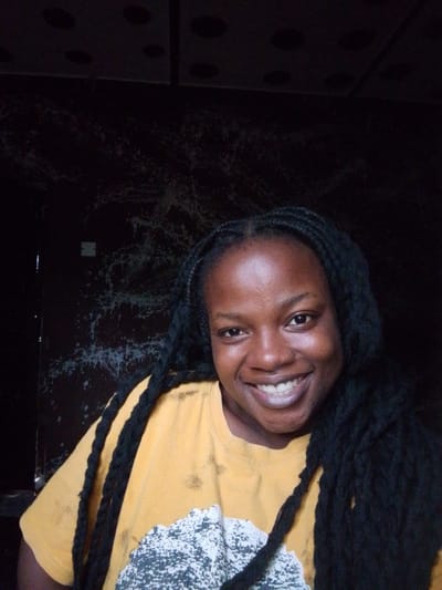 A young woman with dark skin and black braided hair smiles at the camera in a self-portrait. She is wearing a yellow t-shirt with a black and white speckled design on the front. The background is dark and appears to be a wall with some white splatters. The image is dimly lit, with the woman's face and upper body being the brightest points. The location is identified as Evbogida, Nigeria.