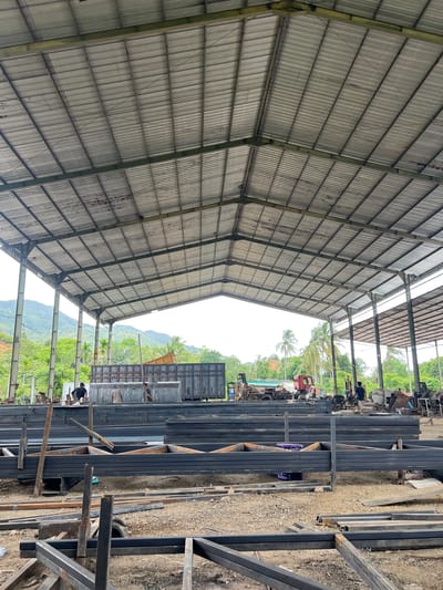 The image depicts an outdoor industrial setting, likely a steel fabrication yard or construction site, located near Lambarobiluy, Indonesia. The scene is dominated by a large, open-sided metal-roofed structure with a corrugated metal roof supported by exposed metal beams and columns. Piles of dark metal beams and structural components are stacked in the foreground and middle ground, occupying much of the ground space.
In the middle ground, there are several large metal containers or truck beds, some appearing to be loaded with materials. A red truck with a yellow bed is visible behind these containers. A few individuals are present, appearing to be workers. One person in a dark t-shirt is standing near the containers, while two others are visible standing on top of or near the containers. Another person in a dark t-shirt is walking in the right-middle ground.
The background reveals a lush, tropical landscape with dense green vegetation, palm trees, and rolling hills or mountains under a cloudy sky. The overall atmosphere suggests an active worksite in a rural or semi-rural Indonesian environment. The ground is covered with dirt, gravel, and scattered debris.