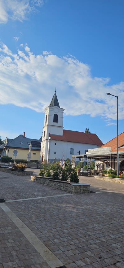 This is a photograph taken outdoors during the daytime, likely in the late afternoon given the shadows. The sky is a clear blue with scattered white clouds. The image features a prominent white church tower with a dark, pointed roof and a small cross at the apex. Adjacent to the church is a building with a red tiled roof, appearing to be a shop or restaurant, indicated by outdoor seating and plants. A sign visible on this building reads "Mi Kis Boltunk". The ground in the foreground is paved with a mosaic of light brown and grey rectangular tiles, suggesting a public area like a square or street. There are some trees and bushes visible, adding greenery to the scene. The overall atmosphere appears peaceful and sunny.