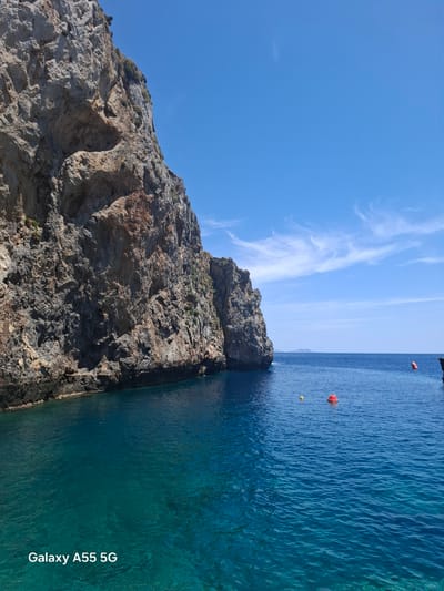 The image depicts a coastal scene near Plakias, Greece. A rocky cliff face dominates the left side of the image. The cliff is composed of light-colored rock formations, showing evidence of erosion and geological layering. The cliff drops directly into clear, turquoise water. Several red and yellow buoys are visible in the water, suggesting a designated swimming or boating area. The sky is a clear, light blue with some wispy clouds. The text "Galaxy A55 5G" is present on the bottom right of the image, which could indicate the device used to take the photo.