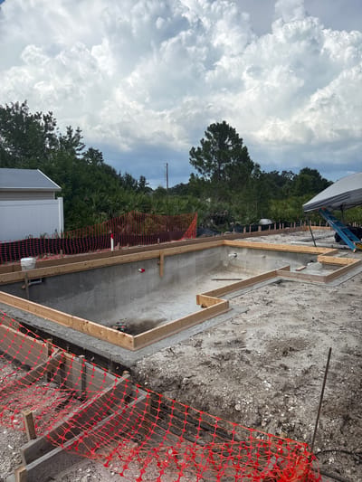 The image shows the construction of a concrete swimming pool in North Port, Florida, United States. The pool is partially constructed, with concrete walls and floor in place, and wooden forms set up around the edges for further concrete pouring. Construction materials, including rebar and tools, are visible on the ground. Orange safety netting is used to cordon off areas of the construction site. In the background, there is a residential building, lush green vegetation including palm trees, and a cloudy sky. A white tent with a ladder underneath is set up on the right side of the frame.