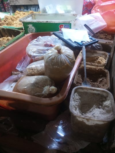 A market stall displays containers of various spices and ingredients. Bags of granular material are packed in plastic, some tied at the top. Several square containers hold powdered substances in shades of brown and grey. A calculator rests on top, next to a spoon. Other items, including additional containers of food, are visible in the background. The lighting is dim, creating shadows that obscure details.