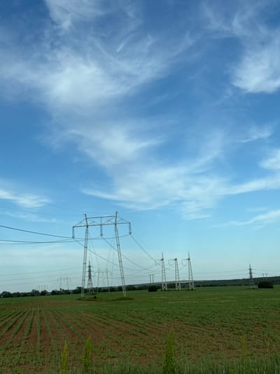 The image depicts a rural landscape in Brestovets, Bulgaria. A green field with rows of crops stretches into the distance, bordering a line of trees. Overhead power lines and transmission towers run along the edge of the field. The sky is blue with scattered white clouds. The image is rotated and tilted.