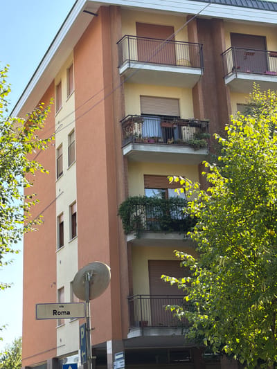 The image shows a view of an apartment building in Vanzaghello, Italy. The building has a light beige facade with orange accents. It features multiple balconies with black railings, some of which are adorned with plants. A street sign indicating "via Roma" is visible in the foreground, along with a blue directional sign. Green trees with dense foliage are present on the left and right sides of the frame, partially obscuring the building. The sky is clear and blue. No people are visible in the image.