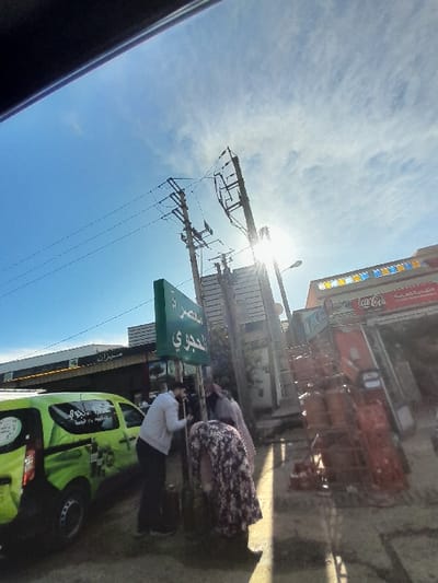 The image captures a sunlit street scene in Oued Jdida, Morocco, under a bright blue sky with wispy white clouds. The sun, positioned low and to the right, produces significant lens flare and casts long shadows towards the left.
On the left, a bright green commercial van featuring white Arabic text and graphics, including a prominent "DELI" logo, is parked. Adjacent to the van stands a green rectangular sign displaying white Arabic text, specifically "مسحري الحجري". Three individuals are gathered near this sign. One male, wearing a light grey long-sleeved shirt and dark trousers, is leaning forward, his back largely to the viewer, engaged with something near the sign's base. To his right, a female clad in a floral-patterned dress or abaya is significantly bent over, facing away from the viewer, appearing to manipulate an object on the ground. A third person, partially obscured behind the male, is visible with a light-colored headscarf.
The right side of the image features multiple utility poles supporting numerous overhead power lines. A building further right exhibits a prominent red "Coca-Cola" sign, below which several red and brown market trolleys are stacked on the pavement. The ground surface is paved.
